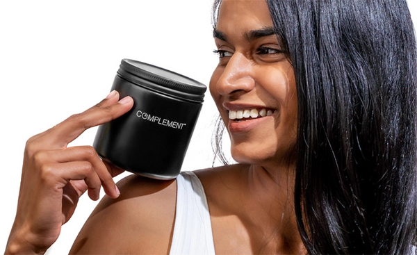 A person with long dark hair smiles while holding a black container labeled COMPLEMENT against a white background. They are wearing a light-colored sleeveless top.