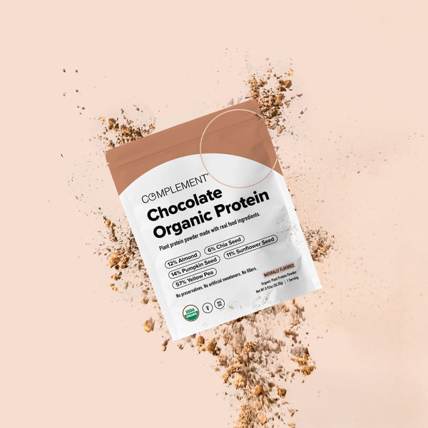A pouch of Complement Chocolate Organic Protein powder is shown with powder scattered around it on a light beige background. The packaging highlights plant protein sources and natural ingredients.