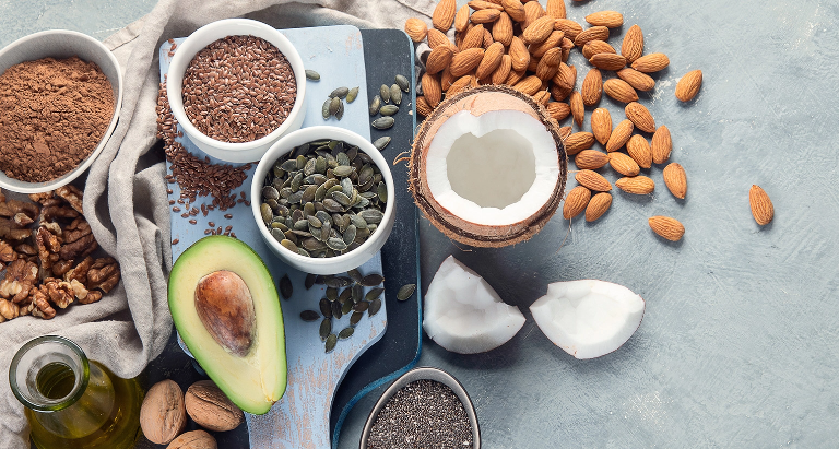 A variety of healthy foods are displayed, including walnuts, flaxseeds, almonds, pumpkin seeds, cocoa powder, and chia seeds. A halved avocado, a broken coconut, and a small bottle of oil are arranged on a light gray surface.