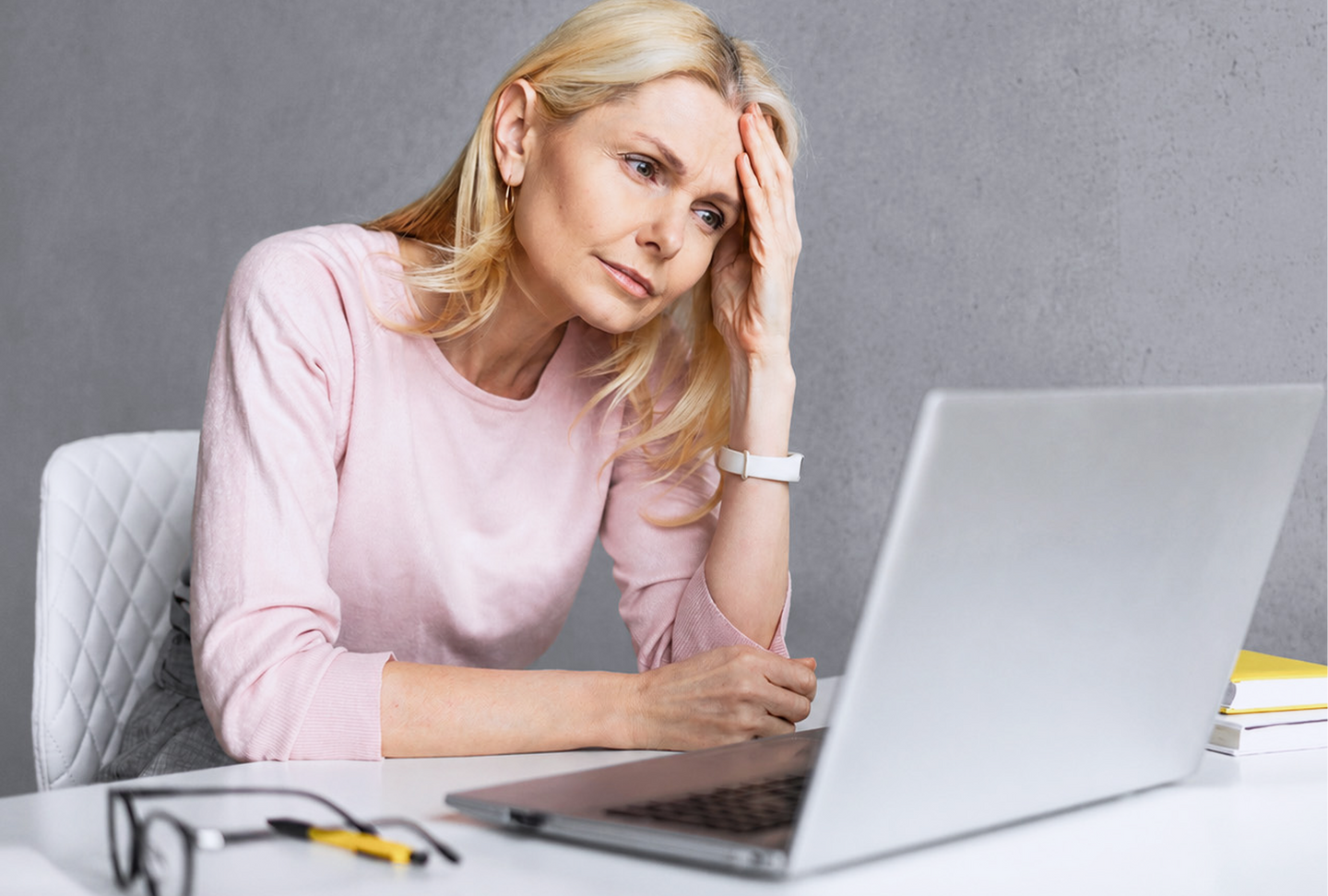 A middle-aged woman with blonde hair sits at a desk looking stressed, resting her head on her hand while staring at a laptop. Glasses, a pen, and a notebook are on the table in front of her.