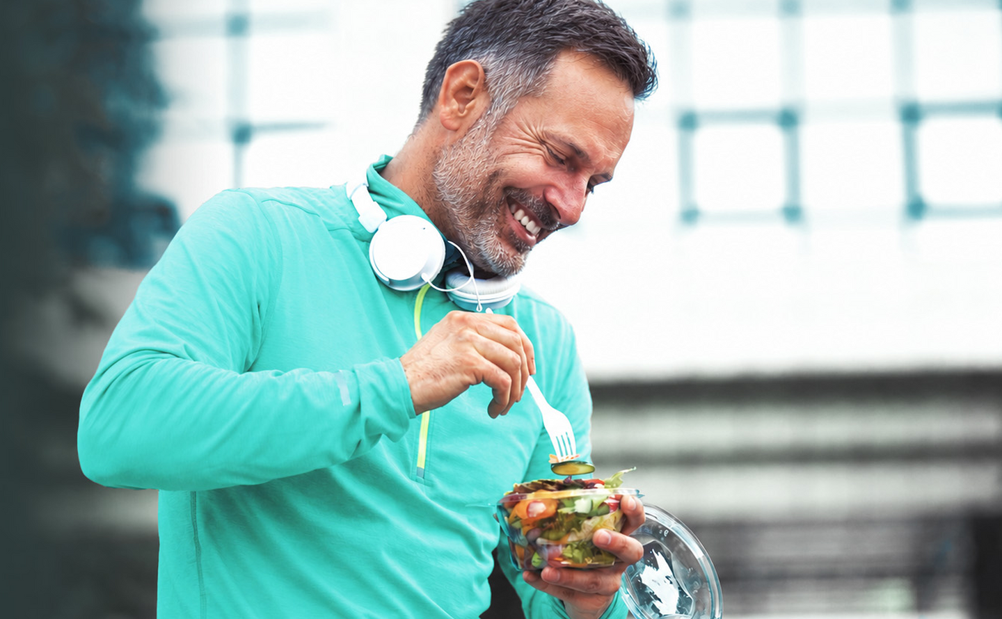 Smiling man in a teal jacket with white headphones around his neck, holding a bowl of salad and eating with a fork outdoors.