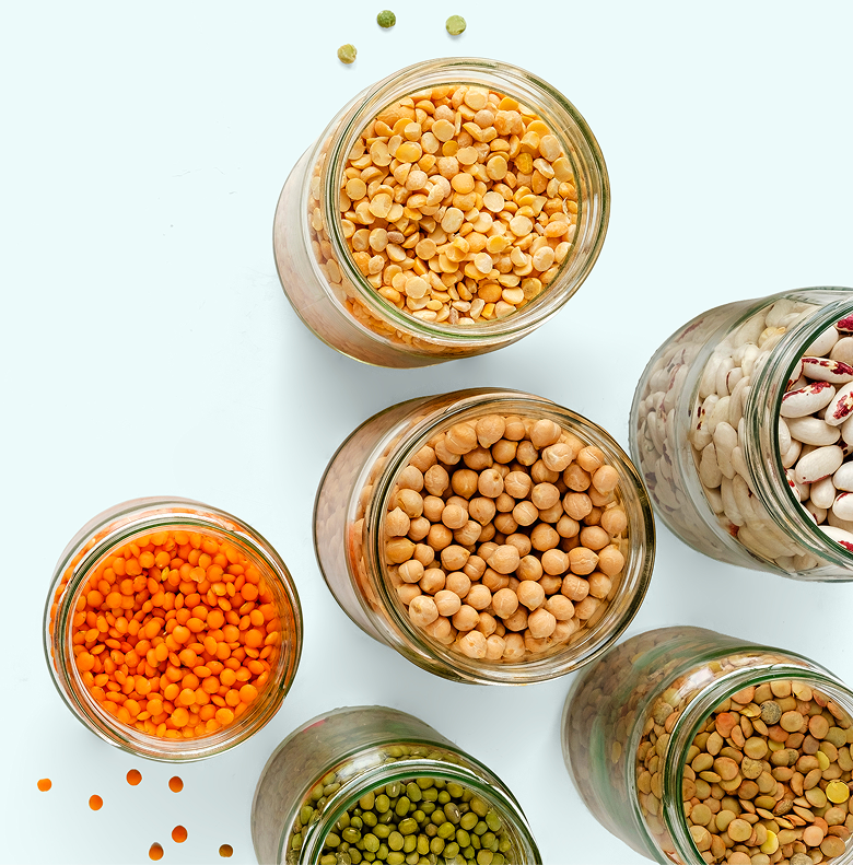 Six glass jars filled with various dried legumes, including lentils, chickpeas, split peas, and beans, are arranged on a white surface. Some legumes are scattered around the jars.