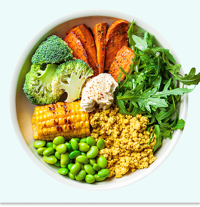 A white bowl filled with broccoli, roasted sweet potato, arugula, grilled corn, edamame, scrambled tofu, and a dollop of hummus, arranged in colorful sections.