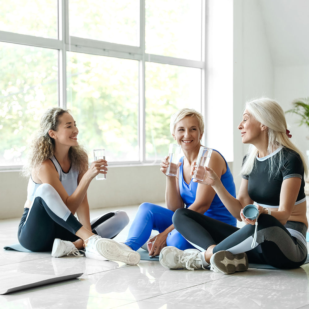 Three women in athletic wear sit on a gym floor, smiling and talking while holding glasses of water, with large windows and greenery visible in the background.