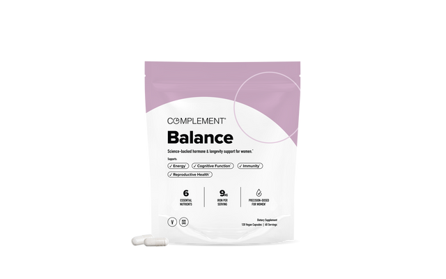 A white and purple pouch labeled Complement Balance stands upright, with two white capsules placed in front. The package highlights benefits like energy, cognitive function, and immunity.