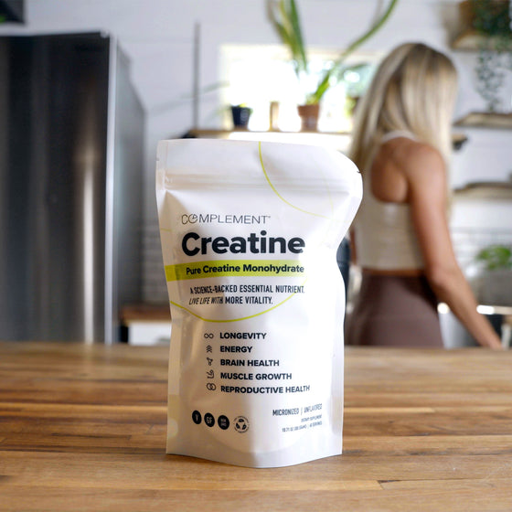 A bag of Complement Creatine Monohydrate sits on a wooden kitchen counter, with text highlighting benefits like longevity, energy, and brain health. A woman stands in the blurred background near kitchen shelves and plants.