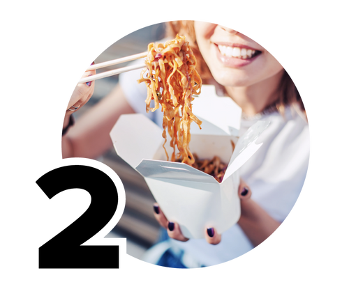 A person is smiling while lifting noodles with chopsticks from a white takeout box. The background is blurred, focusing on the noodles and the persons joyful expression.