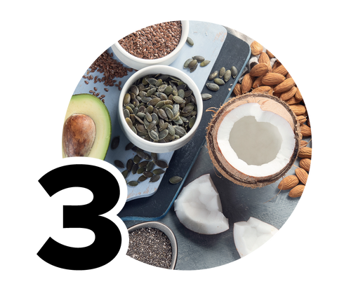 A selection of healthy foods including an avocado half, a bowl of pumpkin seeds, flaxseeds, almonds, chia seeds, and a cracked open coconut, all arranged on a light blue background.