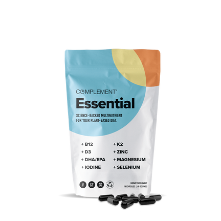A colorful, resealable pouch labeled COMPLEMENT® Essential from Complement stands upright, with black capsules nearby. The package highlights plant-based multivitamin ingredients like B12, D3, K2, DHA/EPA omega-3s, iodine, zinc, magnesium, and selenium.
