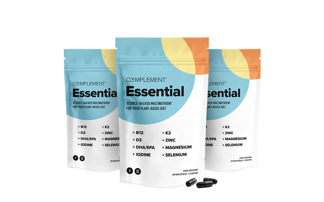 Three Complement Essential supplement pouches with a minimalist blue, orange, and white design are shown, along with two black capsules in front. The packaging lists vitamins and minerals included in the product.