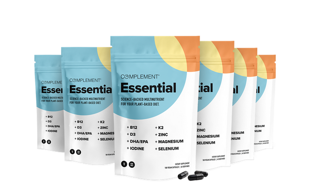 Six standing pouches of Complement Essential multivitamin, with white and blue packaging and a yellow-orange semi-circle design, along with two black capsules shown in front. The label highlights key nutrients for plant-based diets.