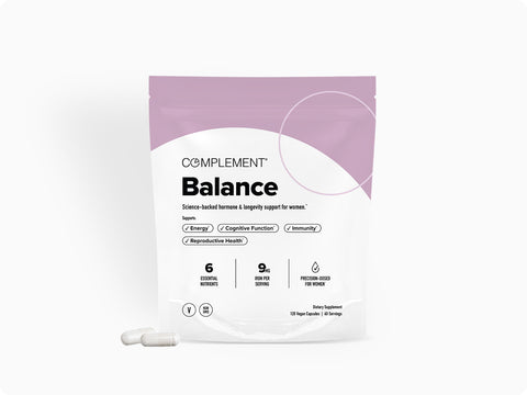 A white and lavender package labeled Complement Balance with supplement details stands upright. Two white capsules are placed in front of the package on a plain white background.