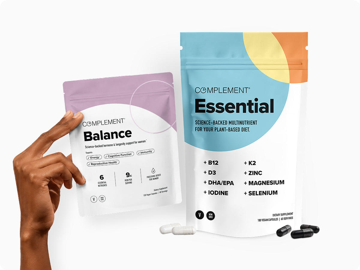 A hand holds a pouch of Complement Balance supplement next to a larger pouch of Complement Essential, with white and black capsules displayed in front.