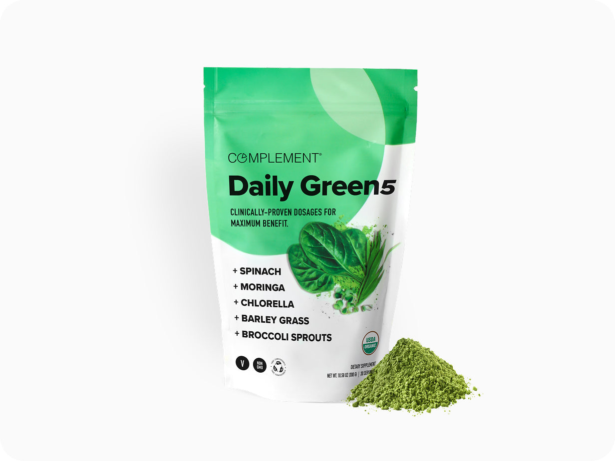 A green and white pouch labeled Complement Daily Green 5 containing a powdered supplement blend. The bag lists spinach, moringa, chlorella, barley grass, and broccoli sprouts. A small heap of green powder is beside the pouch.