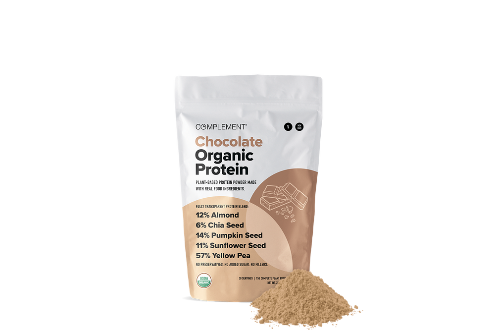 A bag labeled Complement Chocolate Organic Protein with powder spilled in front. The bag highlights plant-based protein with almond, chia, pumpkin, sunflower seeds, and yellow pea.
