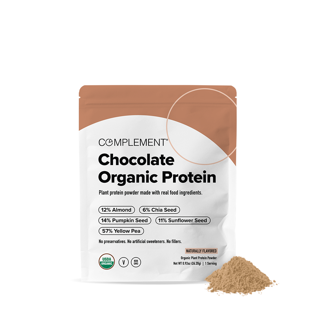 A bag of Complement Chocolate Organic Protein powder with a small mound of powder beside it. The packaging lists almond, chia seed, pumpkin seed, sunflower seed, and yellow pea as ingredients.