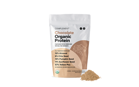 A bag of Complement Chocolate Organic Protein powder stands upright with a small pile of the powder in front. The bag lists ingredients and nutritional percentages, and features a minimalist brown and white design.