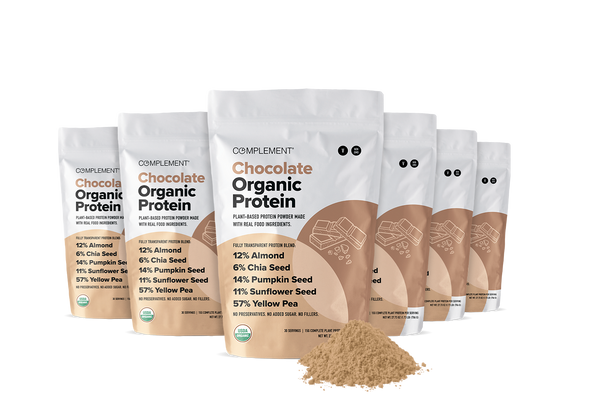 Six standing bags of Complement Chocolate Organic Protein powder are shown, with a small mound of the light brown powder in front. The packaging highlights organic ingredients and plant-based protein.