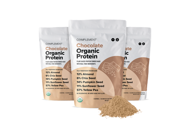 Three bags of Complement Chocolate Organic Protein powder stand upright, showing nutrition details. A mound of chocolate protein powder is in front of the bags on a white background.