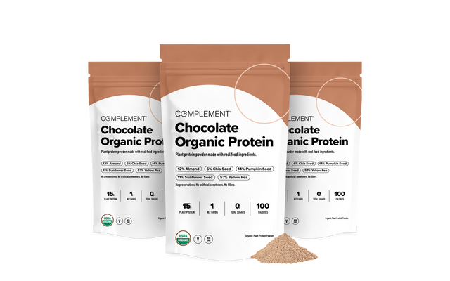 Three bags of Complement Chocolate Organic Protein powder are shown, standing upright, with some powder spilled in front. The packaging highlights plant-based protein and ingredients like chia seeds and pumpkin seeds.