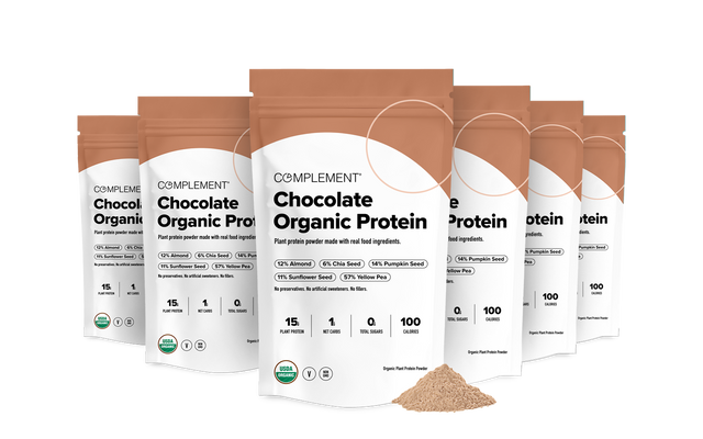 Seven bags of Complement Chocolate Organic Protein powder are arranged in rows, with protein powder piled in front. The packaging highlights plant-based ingredients, nutritional facts, and an organic label.