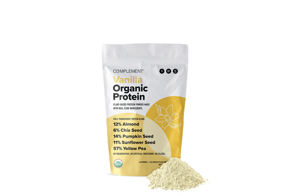 A white and yellow bag labeled Complement Vanilla Organic Protein stands upright with a small mound of light-colored protein powder in front. The bag lists plant-based ingredients like almond, chia seed, pumpkin seed, sunflower seed, and yellow pea.