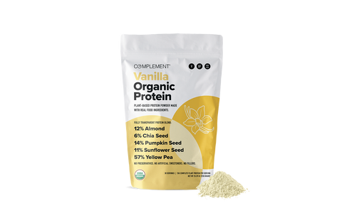 A white and yellow bag labeled Complement Vanilla Organic Protein stands upright with a small pile of protein powder in front. Key ingredients and percentages are listed on the bag, including almond, chia seed, pumpkin seed, sunflower seed, and yellow pea.