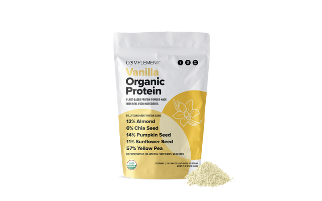 A white and yellow bag labeled Complement Vanilla Organic Protein stands upright with a small pile of protein powder in front. Key ingredients and percentages are listed on the bag, including almond, chia seed, pumpkin seed, sunflower seed, and yellow pea.