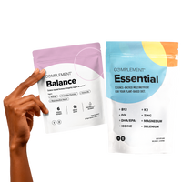 A hand holds two supplement packets: one pink and white labeled Balance and one blue, orange, and white labeled Essential. Both are from Complement and designed for plant-based diets.