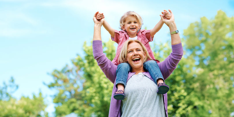 A smiling older woman carries a young girl on her shoulders outdoors, both holding hands and laughing, with green trees and a blue sky in the background.