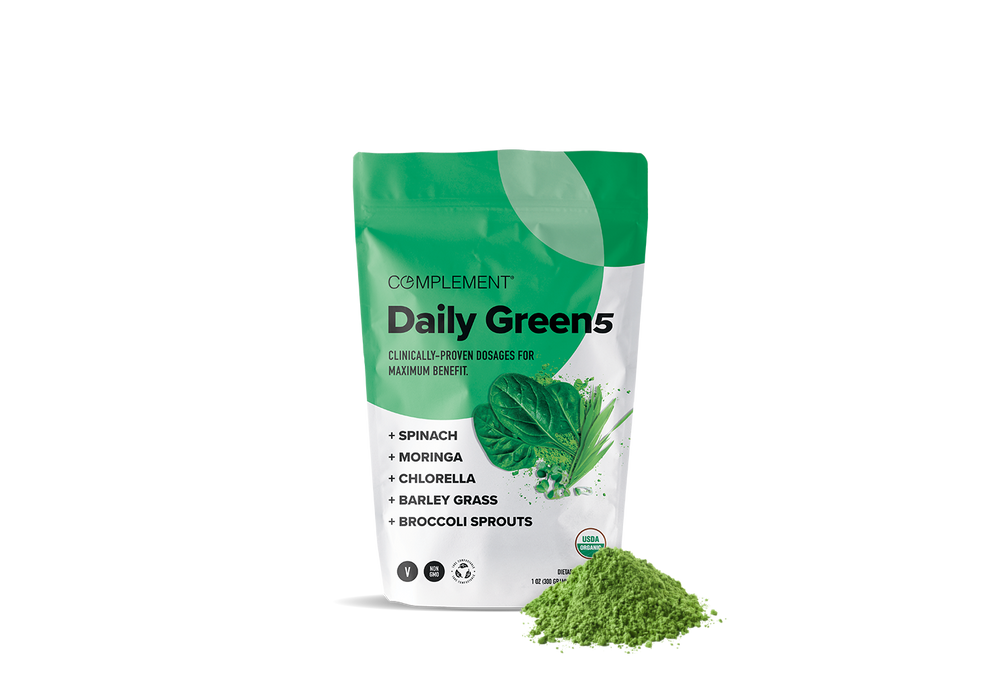 A green and white pouch labeled Complement Daily Green5 stands upright with a heap of green powder in front. The packaging lists ingredients: spinach, moringa, chlorella, barley grass, and broccoli sprouts.