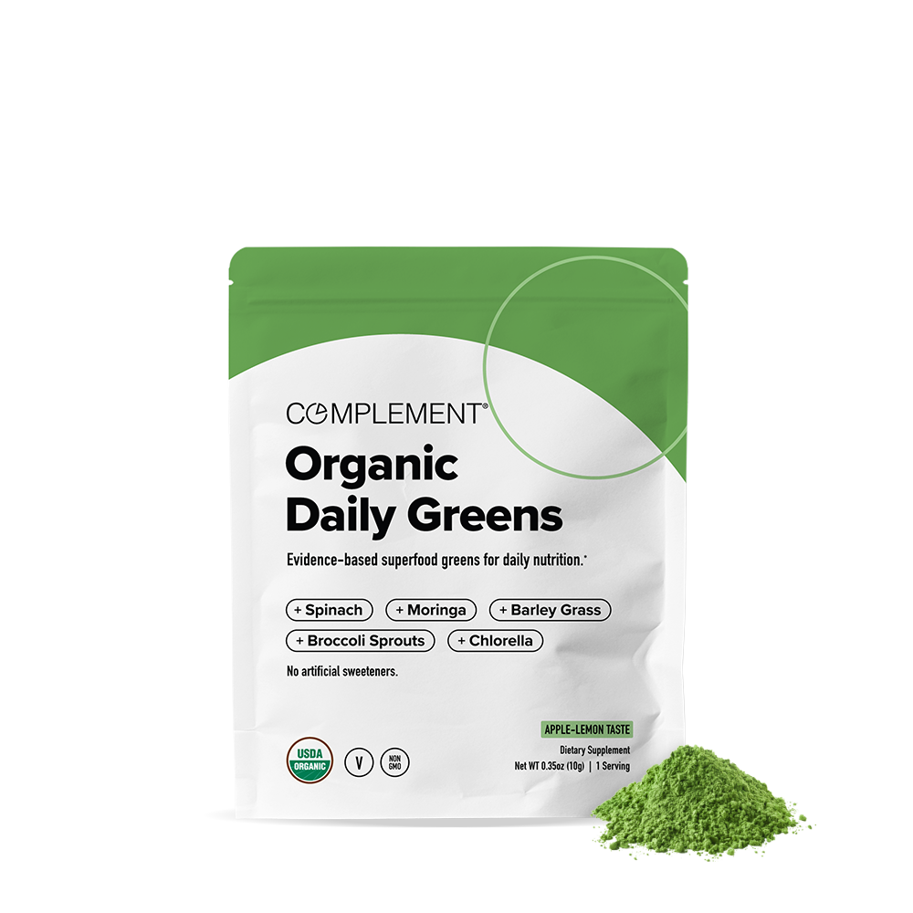 A white and green pouch labeled Complement Organic Daily Greens features ingredients like spinach, moringa, barley grass, broccoli sprouts, and chlorella. A small pile of green powder is shown in front of the pouch.