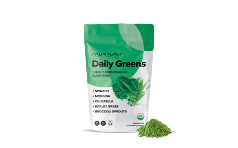 A green and white pouch labeled Complement Daily Green 5 stands next to a small pile of green powder. The package lists spinach, moringa, chlorella, barley grass, and broccoli sprouts as ingredients.