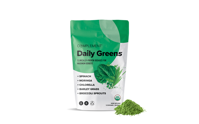 A green and white pouch labeled Complement Daily Green 5 stands next to a small pile of green powder. The package lists spinach, moringa, chlorella, barley grass, and broccoli sprouts as ingredients.