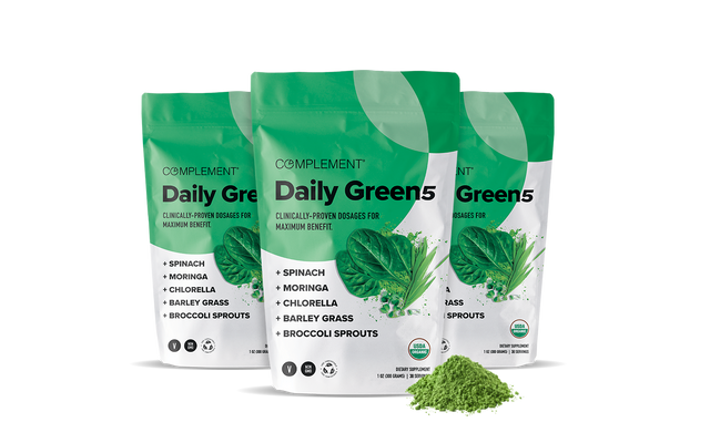Three green and white pouches labeled Complement Daily Green 5 are shown, featuring images of leafy greens. Ingredients listed include spinach, moringa, chlorella, barley grass, and broccoli sprouts; green powder is scattered in front.