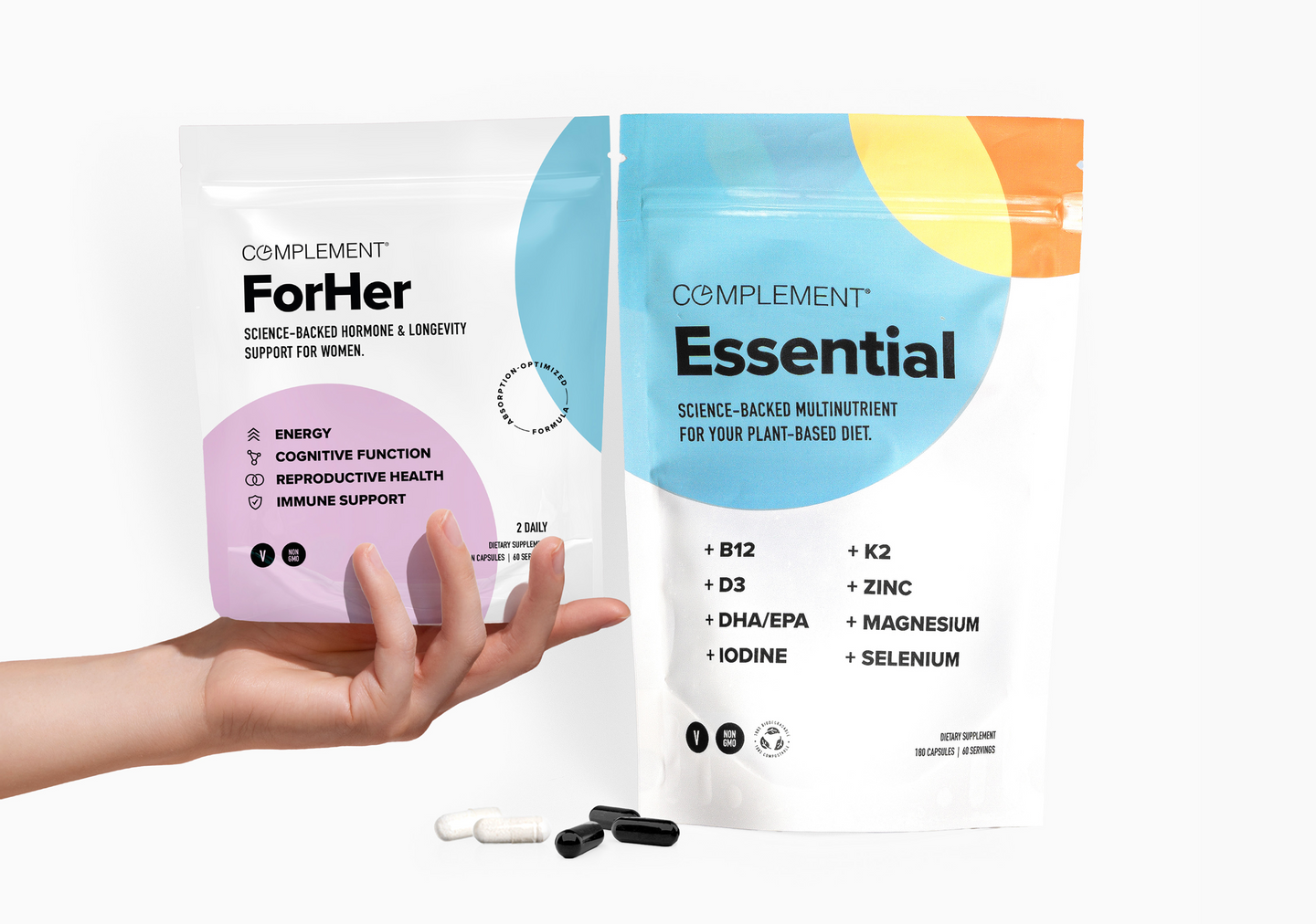 A hand holds a white and purple Complement For Her supplement pouch beside a larger Complement Essential supplement pouch. Both pouches list vitamins and nutrients. Three capsules and one white tablet are on the surface below.