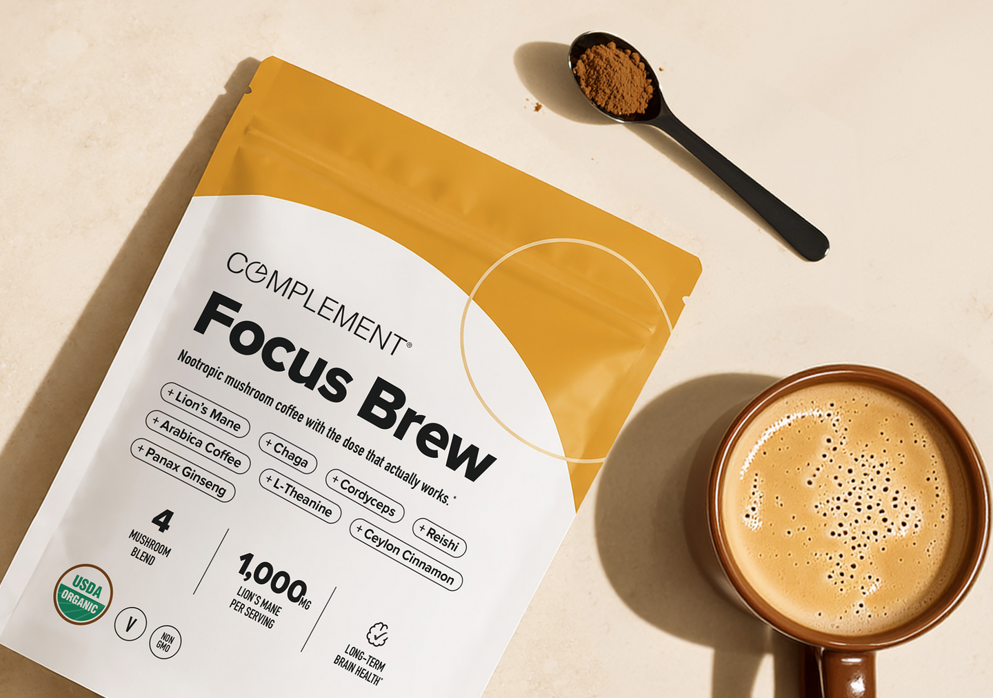 A package of Complement Focus Brew coffee is shown next to a cup of coffee and a black spoon with powder, all set on a light beige surface.