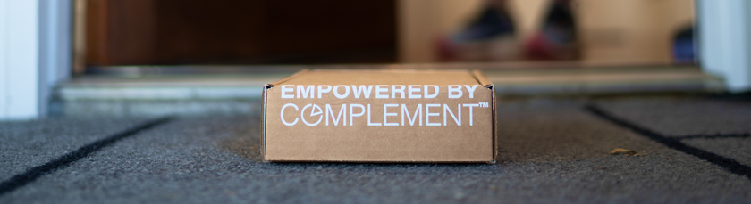 A cardboard box labeled Empowered by Complement sits on a doormat at an open doorway, with a pair of blurred shoes visible in the background.
