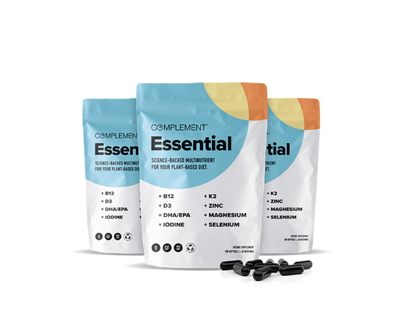 Three pouches of Complement Essential multinutrient supplements are displayed. The packaging is white with colorful shapes, featuring nutrients like B12, D3, DHA/EPA, K2, zinc, and more. Black capsules are scattered in front of the pouches.