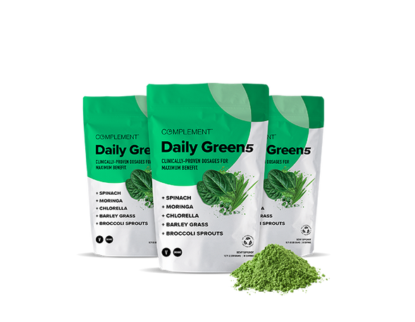 Three green and white pouches of Complement Daily Green5 dietary supplement powder are displayed, featuring spinach, moringa, chlorella, barley grass, and broccoli sprouts. A small pile of the green powder is in front of the pouches.