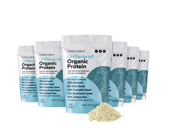 Bags of Complement Unflavored Organic Protein are arranged in a staggered row. The packaging highlights plant-based protein sources, including almond, chia seed, pumpkin seed, sunflower seed, and yellow pea. Powder is spilled in front of the bags.
