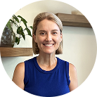 A woman with short blonde hair wearing a sleeveless blue top smiles at the camera. There is a plant and wooden shelves in the background.