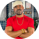 A man wearing a red shirt, beige cap, gold chain, and bracelets stands with arms crossed in a modern kitchen with blue tiles and spice jars on the shelves behind him.