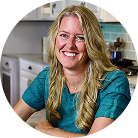 A smiling woman with long blonde hair and a teal top sits at a kitchen counter with white cabinets and teal backsplash tiles in the background.