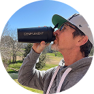 A man wearing a gray hoodie, glasses, and a cap drinks from a black bottle labeled COMPLIMENT outdoors on a sunny day with green grass and trees in the background.