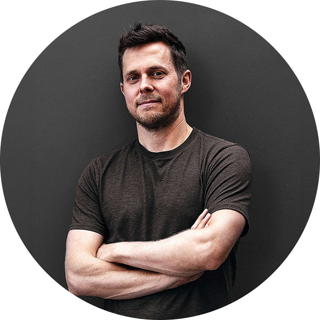 A man with short dark hair and a trimmed beard stands against a dark gray wall, arms crossed, wearing a dark t-shirt and smiling slightly.
