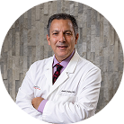 A middle-aged man wearing a white lab coat and a dress shirt with a tie stands in front of a textured, neutral-colored wall, smiling confidently at the camera.