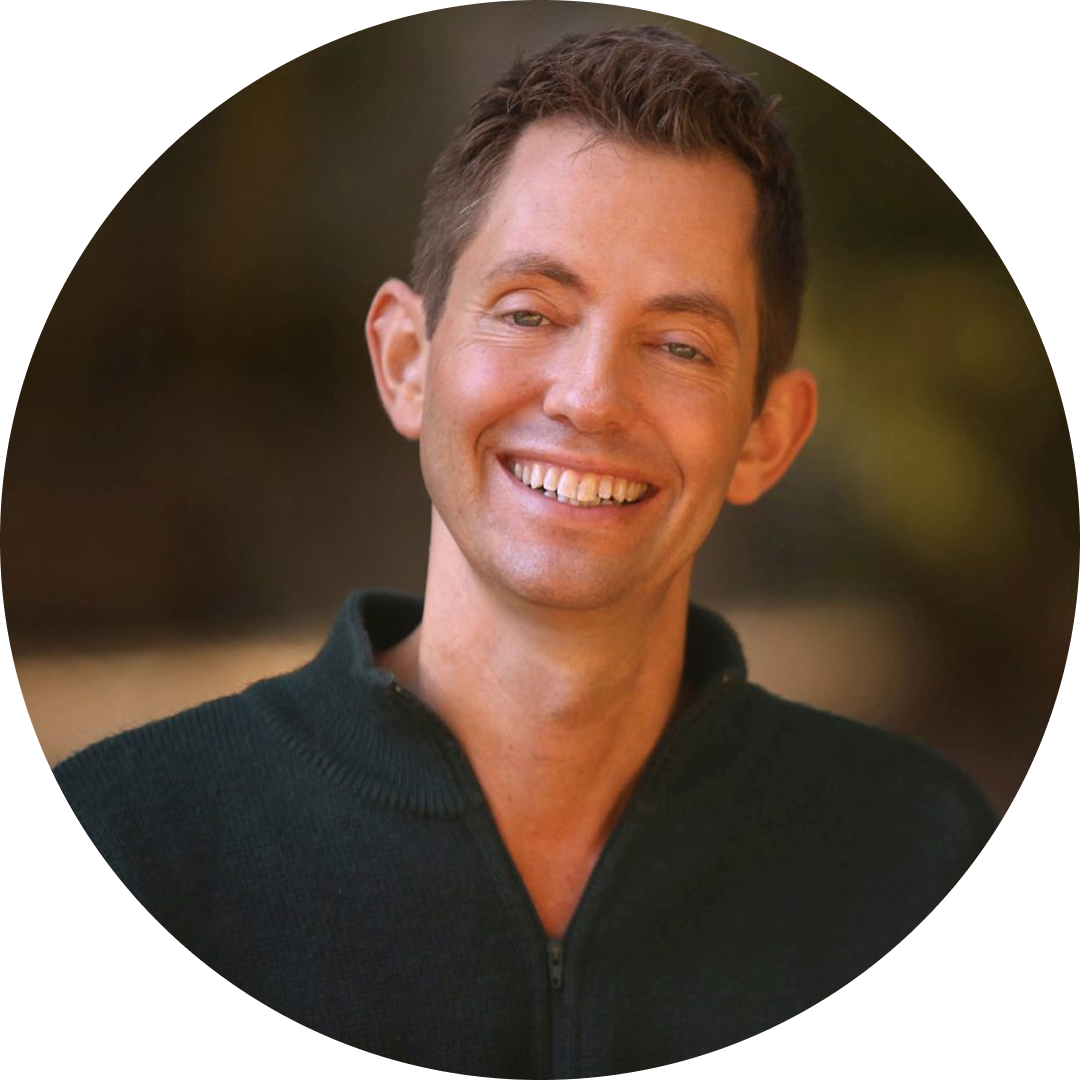 A smiling man with short brown hair wearing a dark zip-up sweater, photographed against a softly blurred outdoor background.