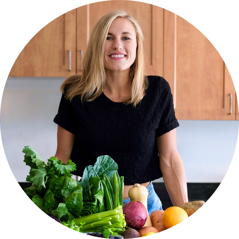 A woman with blonde hair and a black shirt smiles while standing in a kitchen behind a table filled with assorted fresh vegetables and fruits.