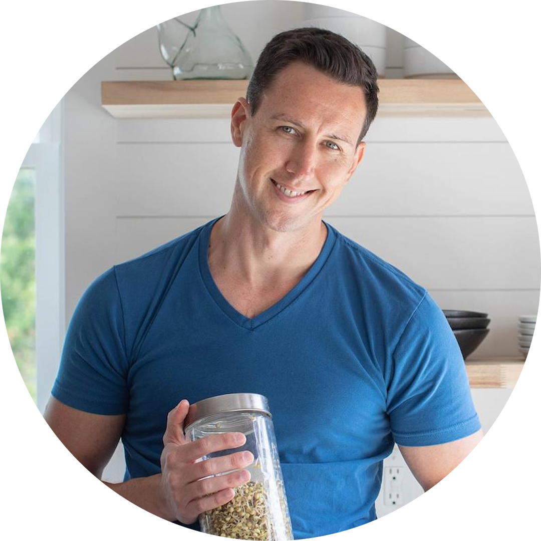 A man in a blue t-shirt smiles while holding a jar of dried food in a bright, modern kitchen with white walls and wooden shelves.